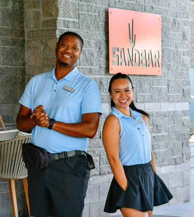 Two people in matching light blue shirts standing in front of a stone wall with a 'SANDBAR' sign.